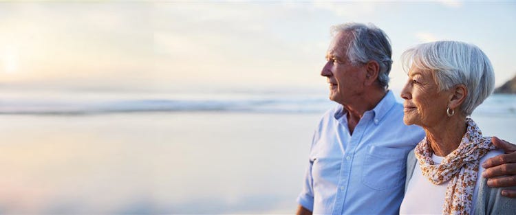 Actor portrayal of an older man and woman standing side by side looking out toward the ocean shoreline