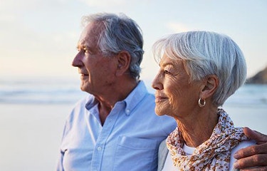 Actor portrayal of an older man and woman standing side by side looking out toward the ocean shoreline