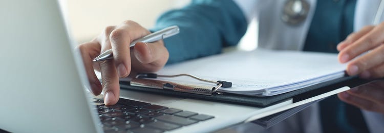 A healthcare professional's hand holding a pen and typing on a computer