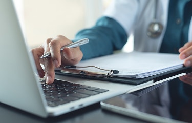 A healthcare professional's hand holding a pen and typing on a computer
